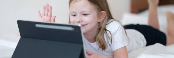 Mała dziewczynka leżąca na łóżku i machająca ręką na ekranie tabletu. Aplikacje dla dzieci mobilna koncepcja - Zdjęcie, obraz