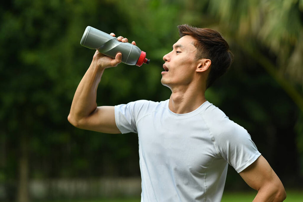 Uomo che beve acqua dalla bottiglia, riposandosi dopo aver fatto jogging al parco la sera. Stile di vita sano e concetto di fitness.  - Foto, immagini