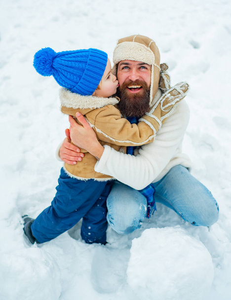 "Zoon knuffelt zijn vader op wintervakantie. Pa en zoontje spelen buiten samen. Gelukkig kind spelen met sneeuwbal tegen witte winter achtergrond." - Foto, afbeelding