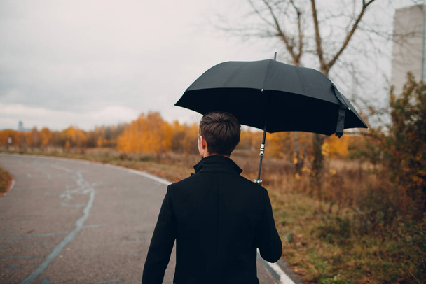 "Młody człowiek w czarnym płaszczu, spacery w deszczu z parasolem" - Zdjęcie, obraz