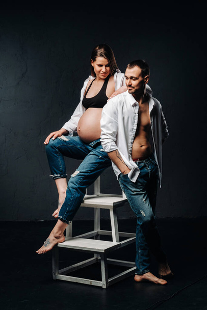 "těhotná žena a muž v bílé košili a džíny v ateliéru na černém pozadí" - Fotografie, Obrázek