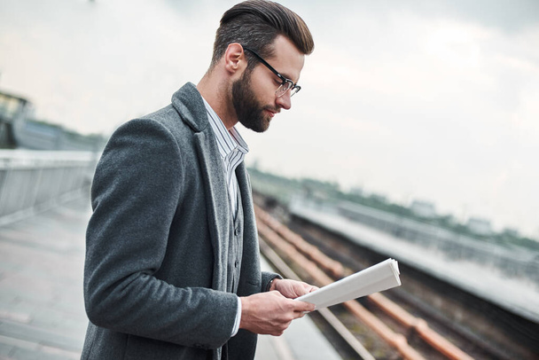 Business trip. Young businessman standing near railway reading newspaper close-up - Foto, afbeelding