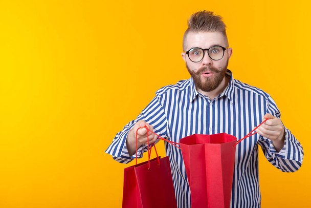 Verrast vrolijke jonge knappe man met een bril en een baard kijkt naar de pakketten om te winkelen, staand op een gele achtergrond. Het concept van succesvolle aankopen. Advertentieruimte. - Foto, afbeelding