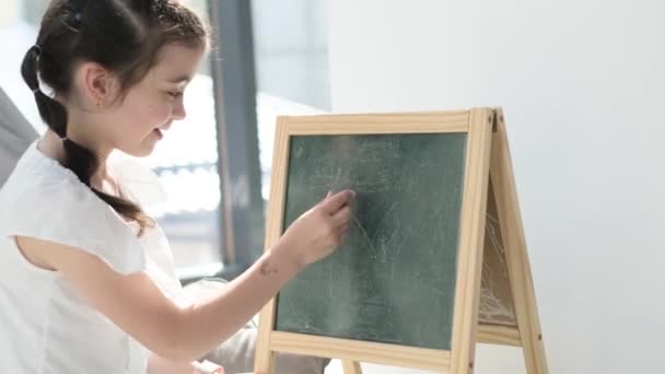 Een klein meisje schrijft met krijt op een schoolbord. Basisschool voor kinderen, slim kind studeert - Video