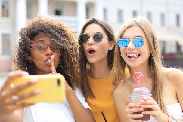 Tres chicas hipster cool de moda, amigos beber cóctel y tomar selfie en el fondo de la ciudad
. - Foto, Imagen