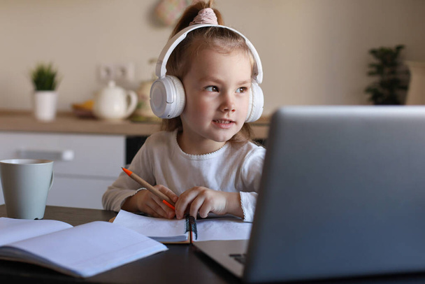 Kulaklıkla gülümseyen küçük bir kız evde dizüstü bilgisayar kullanıyor, kulaklıklı şirin mutlu bir çocuk internet dersi alıyor ya da bilgisayar dersi alıyor.. - Fotoğraf, Görsel