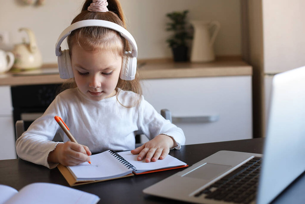 Dziewczynka w słuchawkach siedzieć przy biurku pisząc w notebooku studiując online robić ćwiczenia w domu, małe dziecko pismo ręczne przygotować pracę domową na kwarantannie, mieć klasy internetowej lub lekcji w pomieszczeniach. - Zdjęcie, obraz