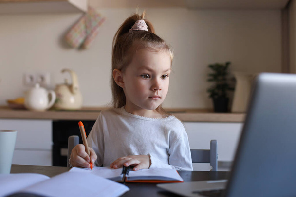 Σοβαρό μικρό κορίτσι handwrite μελέτη σε απευθείας σύνδεση χρησιμοποιώντας το φορητό υπολογιστή στο σπίτι, χαριτωμένο χαρούμενο μικρό παιδί να λάβει Διαδίκτυο μάθημα web ή τάξη στον υπολογιστή. - Φωτογραφία, εικόνα