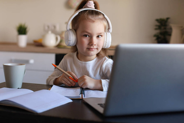 Uśmiechnięta dziewczynka w słuchawki nauka pisma ręcznego online za pomocą laptopa w domu, słodkie szczęśliwe małe dziecko w słuchawki wziąć lekcje Internetu lub klasy na PC. - Zdjęcie, obraz