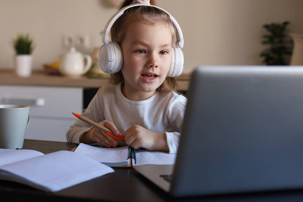 Sonriente niña en los auriculares estudio de escritura a mano en línea utilizando el ordenador portátil en casa, lindo niño pequeño feliz en los auriculares tomar lección web de Internet o clase en PC. - Foto, imagen