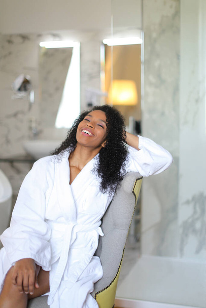 Young happy black girl wearing bathrobe and sitting in bathrom. - Фото, изображение