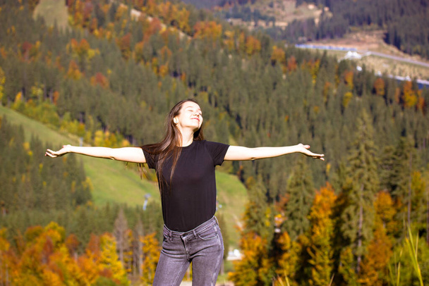 Счастливая белая женщина, девушка в джинсах, черная футболка, стоящая с распростертыми руками на фоне осеннего пейзажа высоко в горах осенним днем. Путешествие осенью, осенью. Буковель-Украина. - Фото, изображение