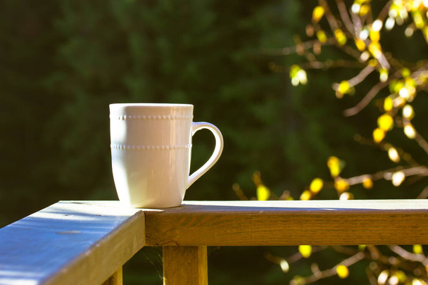 A white cup of tea, coffee hot beverage stands on wooden railing on terrace, balcony against autumnal forest. Relaxation in nature. Good autumn sunny morning, day concept. Fall season sunlight. - Photo, Image