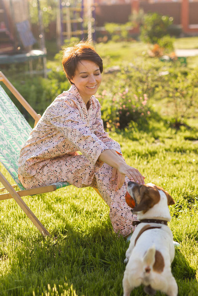 Jeune femme en pyjama repose sur une chaise sur une pelouse verte le jour ensoleillé de l'été - concept de vie de village et de campagne - Photo, image