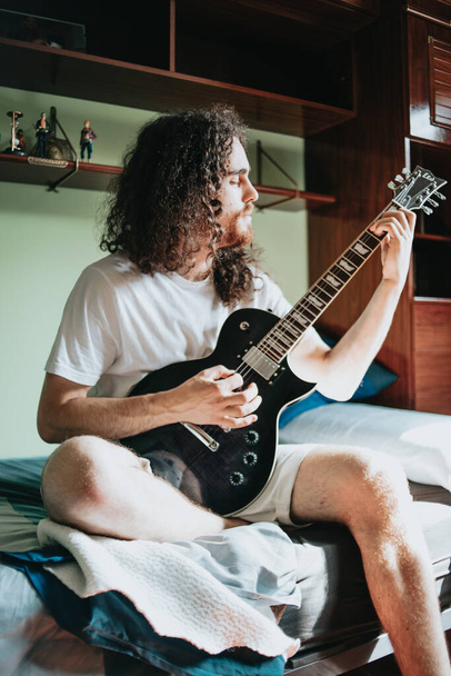 "Молодой человек с длинными волосами играет на электрогитаре в своей комнате. Белая рубашка сидит на уютной кровати в спальне и думает о чем-то. Образ концепции музыкального образования." - Фото, изображение