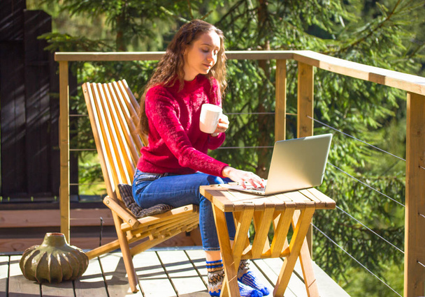 Młoda piękna kobieta z długimi włosami pracuje zdalnie na laptopie siedząc na pięknym tarasie, balkonie w wiejskim domu w lesie iglastym, lesie. Stylowa dziewczyna z białą filiżanką kawy relaksuje. - Zdjęcie, obraz