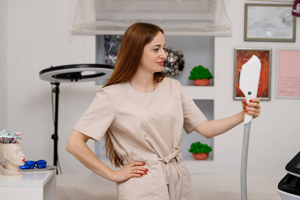Beautician holding ultrasound device for face and skin lifting procedure and hair removal. Young woman cosmetologist using modern cosmetology equipment in clinic. - Φωτογραφία, εικόνα