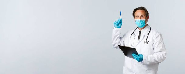 Ковид-19, профилактика вируса, медицинские работники и концепция вакцинации. Задумчивый возбужденный врач-мужчина в медицинской маске и перчатках, держите планшет, поднимая ручку на жест эврики, есть решение - Фото, изображение