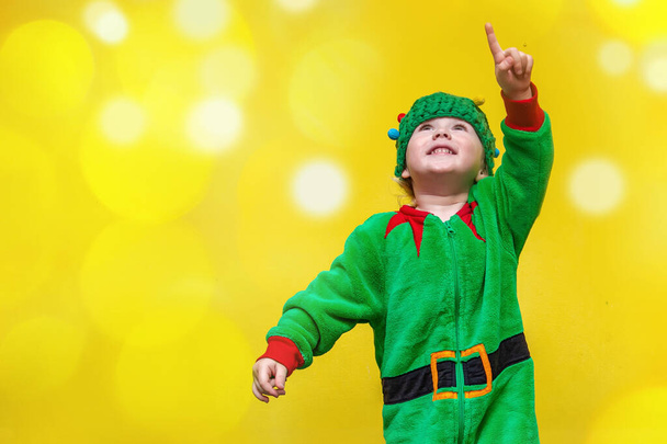 klein meisje in elf kostuum en kerstboom-vormige hoed galoppeert op gele achtergrond wijst vinger omhoog. Reclamebanner. Een plaats voor tekst. - Foto, afbeelding