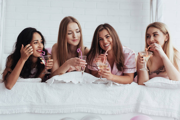 Випив хороші коктейлі. Радісні дівчата в нічному одязі лежать на ліжку в білій кімнаті і святкують - Фото, зображення