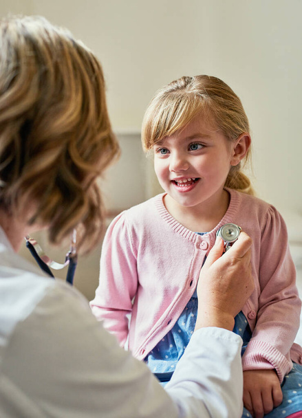Gesundes Herz gleich glückliches Herz. Aufnahme eines Arztes, der ein kleines Mädchen mit einem Stethoskop in ihrem Sprechzimmer untersucht. - Foto, Bild