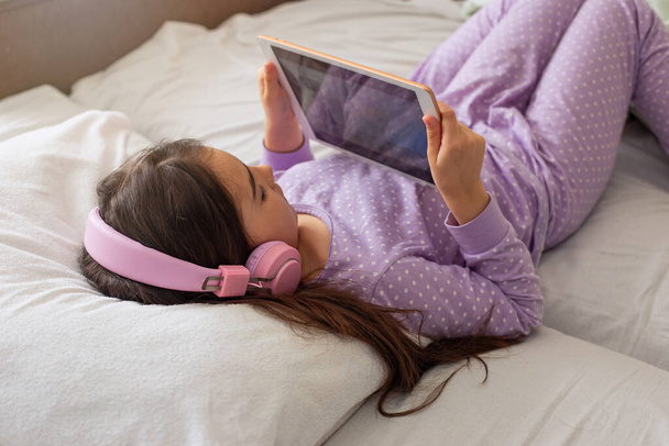 "Ein kleines Mädchen im lila Schlafanzug liegt auf einem weißen Bett auf dem Rücken, hält ein pinkfarbenes digitales Tablet in der Hand." - Foto, Bild