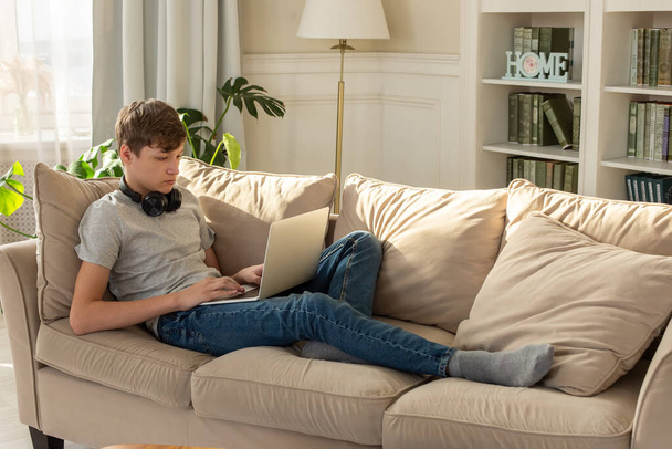 "Nastolatek siedzi na beżowej kanapie, w pokoju z rośliną, mając czarne słuchawki na szyi, zagląda do laptopa.." - Zdjęcie, obraz