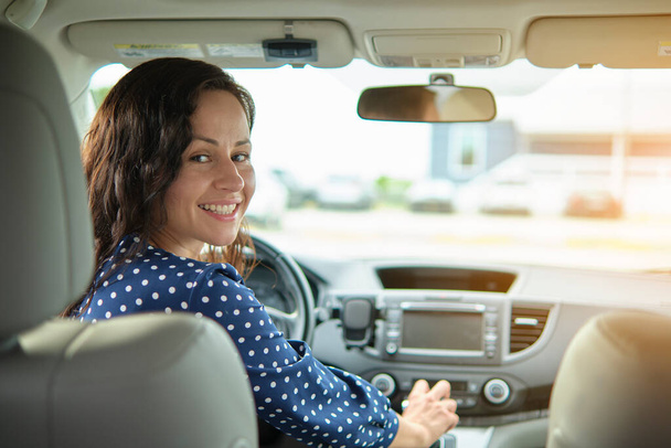 Πορτρέτο της ελκυστική νεαρή γυναίκα σε casual φόρεμα κοιτάζοντας πάνω από τον ώμο της, ενώ κάθεται πίσω από τιμόνι οδήγηση αυτοκινήτου. - Φωτογραφία, εικόνα