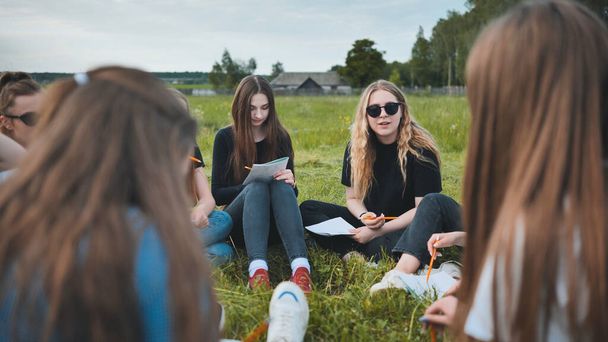 "Група студентів-жінок сидить у колі на лузі для колективної роботи з блокнотами
." - Фото, зображення