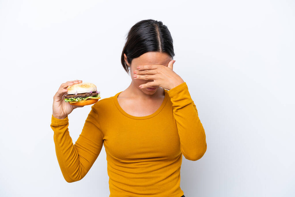 Junge hispanische Frau hält einen Burger isoliert auf weißem Hintergrund mit müdem und kranken Gesichtsausdruck - Foto, Bild