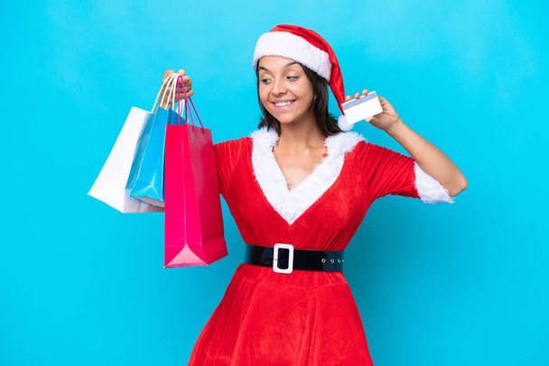 Anne Noel gibi giyinmiş genç İspanyol kadın mavi arka planda izole edilmiş. Elinde alışveriş çantaları ve bir kredi kartı var. - Fotoğraf, Görsel