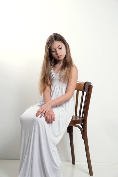 A cute teenage girl in a long white dress with flowing hair sits on a light background on a chair. - Foto, Imagen