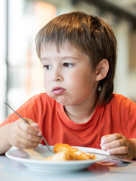 Ο ιδιότροπος με το κόκκινο μπλουζάκι τρώει τηγανίτες με μαχαίρι και πιρούνι. Μικρό αγόρι με αστεία έκφραση στο πρόσωπο. Νόστιμα γλυκά για πρωινό. - Φωτογραφία, εικόνα
