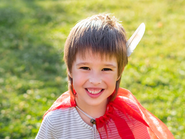 "Retrato de un niño sonriente jugando a ser indio americano. Niño con pluma de pájaro blanco y capa roja. Juego de roles de disfraces. Actividades de ocio al aire libre." - Foto, imagen