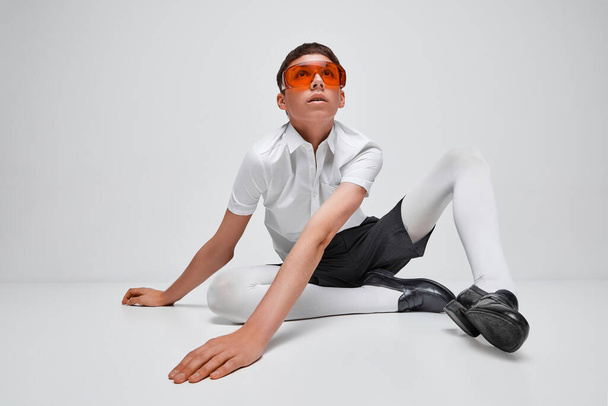 Portret stylowego chłopca pozującego w czarno-białe ubrania z czerwonymi okularami odizolowanymi na szarym tle. Modelowanie. Koncepcja nowoczesnej mody, fotografii artystycznej, stylu, queer, wyjątkowości, reklamy - Zdjęcie, obraz