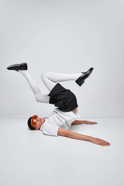 Portrait of stylish boy posing in black and white clothes with red glasses isolated over grey background. Lying on back with legs up. Concept of modern fashion, art photography, style, uniqueness, ad - Zdjęcie, obraz