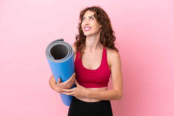 Young sport woman going to yoga classes while holding a mat isolated on pink background thinking an idea while looking up - Photo, image