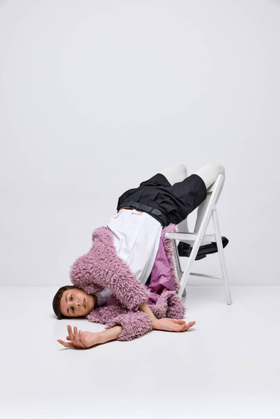 Retrato de niño con estilo en abrigo peludo rosa acostado en silla con las piernas hacia arriba, posando aislado sobre fondo gris. Concepto de moda moderna, fotografía de arte, estilo, queer, singularidad, anuncio - Foto, Imagen