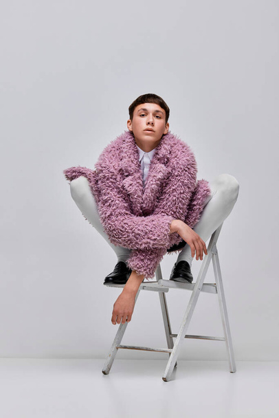 Portrait of stylish boy in pink furry coat posing on chair isolated over grey background. Youth culture. Concept of modern fashion, art photography, style, queer, uniqueness, ad - Фото, изображение
