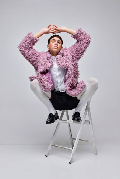 Портрет стильного мальчика в розовом пушистом пальто, позирующем на стуле, изолированном на сером фоне. Молодежная культура. Самовыражение. Современная мода, художественная фотография, стиль, queer, уникальность, реклама - Фото, изображение