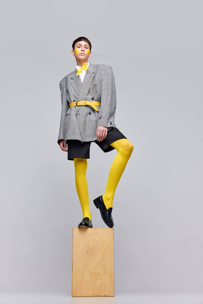 Portrait of stylish boy posing in jacket, blazer and yellow tights and bow isolated over grey background. Statue style. Concept of modern fashion, art photography, style, queer, uniqueness, ad - Fotó, kép