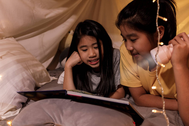 Concept de famille. sœur aînée et sœur lecture livre avec lampe de poche ensemble dans la tente des enfants avant le coucher. Bonne sœur aînée lire livre d'histoire à sa sœur dans la tente drap de lit - Photo, image