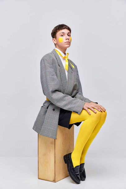Portrait of stylish boy posing in jacket, blazer and yellow tights isolated over grey background. Feminine behavior. LGBT. Concept of modern fashion, art photography, style, queer, uniqueness, ad - Photo, Image