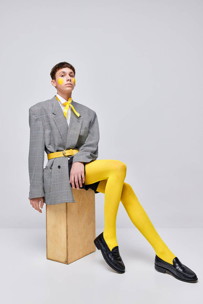Portrait of stylish boy posing in jacket, blazer and yellow tights isolated over grey background. Femininity and lgbt. Concept of modern fashion, art photography, style, queer, uniqueness, ad - Photo, Image