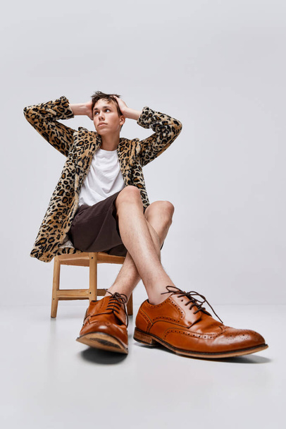 Portret stylowego nastolatka w płaszczu ze zwierzęcym nadrukiem i klasycznych butach brogue, pozującego na szarym tle. Unikalny styl. Koncepcja nowoczesnej mody, fotografii artystycznej, stylu, queer, wyjątkowości, reklamy - Zdjęcie, obraz