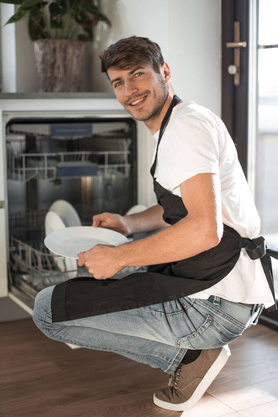 "Yakışıklı genç adam açık bir bulaşık makinesinin yanında oturuyor." - Fotoğraf, Görsel