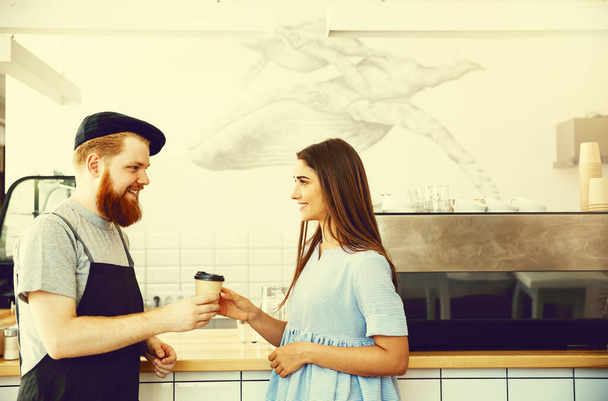 "Кофе Бизнес Концепция - Положительный молодой бородатый мужчина и красивая привлекательная пара бариста в фартуке, глядя в камеру, стоя у барной стойки
" - Фото, изображение