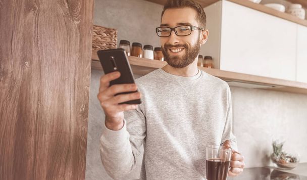 "hombre sonriente con teléfono inteligente y taza de café de pie en su cocina" - Foto, imagen