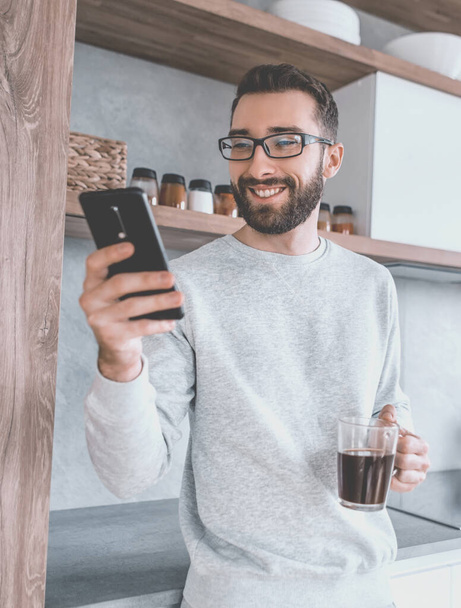"На кухні стоїть усміхнений чоловік із смартфоном та чашкою кави." - Фото, зображення