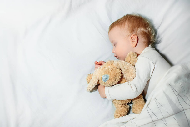 Милая маленькая девочка спит на белом белье в постели. ребенок 1 год спит обнимая мягкую игрушку. Счастливое детство и проблемы со сном у детей - Фото, изображение